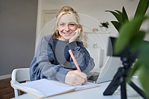 Cute smiling girl sits in a room, writes down notes, doing homework, records video of herself on digital camera, creates