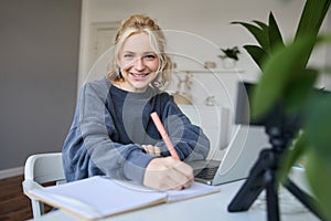 Cute smiling girl sits in a room, writes down notes, doing homework, records video of herself on digital camera, creates