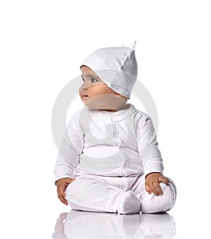 Cute smiling infant baby toddler sits on the floor in white onepiece overall and funny hat with ears and looks aside