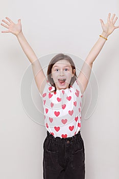 Cute smiling kid girl in white t-shirt with red hearts and stylish black jeans happily raises his hands up on white