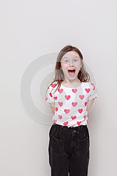 Cute smiling kid girl in white t-shirt with red hearts, stylish black jeans opens his mouth wide on white background