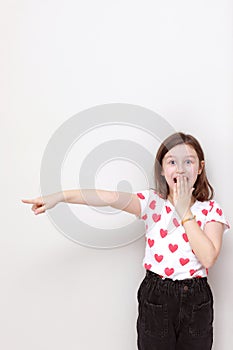 Cute smiling kid girl in white t-shirt with red hearts, stylish black jeans wonderingly pointing her finger looking to