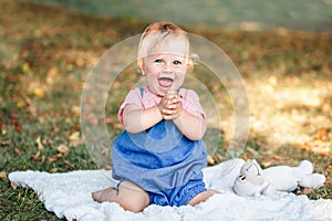 Cute smiling laughing baby girl sitting on white blanket in park outdoors. Adorable excited child toddler playing and having fun