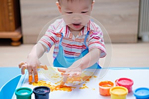 Cute smiling little Asian toddler boy child finger painting with hands and watercolors, kid painting at home
