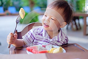 Cute smiling little Asian  toddler boy child sitting in high chair using fork eating pineapple, Healthy snacks & Self feeding conc