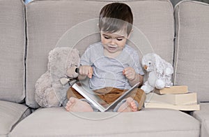 Cute smiling little baby boy reading book sitting on sofa with teddy bear toy and bunny rabbit at home.