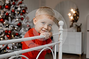 Cute smiling little boy in red sweater sitting on bed holding bed frame with both hands on the background of the Christmas tree.
