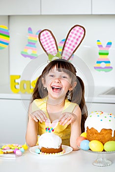 A cute smiling little girl with bunny ears prepares an Easter cake and painted eggs. Religious holiday