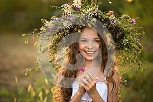 Cute smiling little girl with flower wreath on the meadow at the farm. Portrait of adorable small kid outdoors. Midsummer