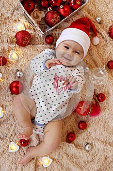 Cute smiling little girl in Santa Claus red hat is playing with wooden toy on a beige plaid with Christmas decorations.