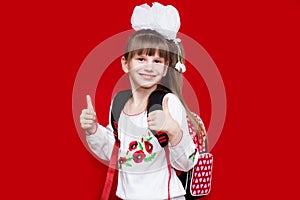 Cute smiling little girl in school uniform and white bows with backpack on red background