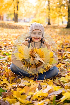 Cute smiling little girl sitting on the fallen leaves in autumn park and holding the bouquet of yellow maple leav