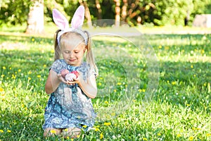 Cute smiling little girl wearing bunny ears on Easter day. Girl sitting on a grass and holding painted Easter eggs