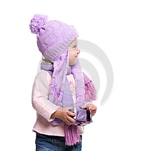 Cute smiling little girl wearing violet knitted scarf and hat, holding christmas gift isolated on white background. Winter clothes