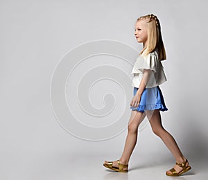 cute smiling little girl in a white blouse with flounces, blue shorts and sandals, walks briskly and looks to the side.