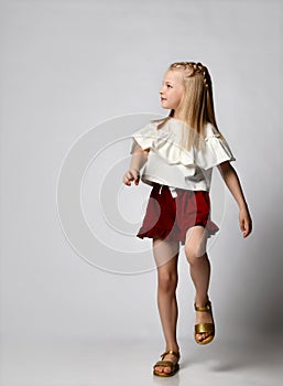 cute smiling little girl in a white blouse with flounces, burgundy shorts and sandals, walks briskly and looks to the