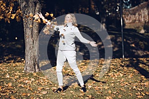 Cute smiling middle-aged woman with long wavy fair hair in grey roll-neck sweater throw up pile of yellow maple leaves.
