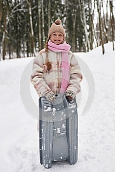 Cute smiling pre-teen girl in winterwear holding toboggan sledge