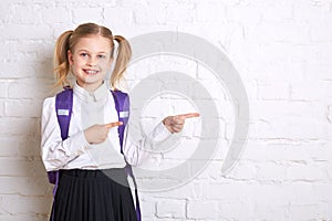 Cute smiling schoolgirl in uniform standing on light background and showing thumbs to the side.