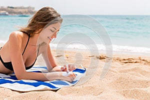 Cute and smiling woman is applying sunscreen on her hand with the finger sunbathing on the towel at the beach