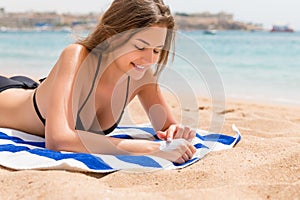 Cute and smiling woman is applying sunscreen on her hand with the finger sunbathing on the towel at the beach