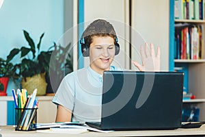 Cute smiling young boy in blue shirt sitting behind desk in his room next to laptop and study. Teenager in earphones waving hand