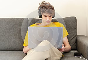 Cute smiling young teenager with laptop and headphones on the couch at home. Boy makes homework with a computer.