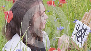 Cute smilling girl painting with black colors on the poppy field. Young woman artist outdoors. Connection with nature