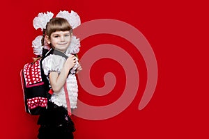 A cute smilng little girl in school uniform and white bows with a backpack on red background. Back to school