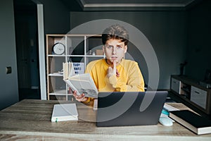 Cute student with a book in his hands sits at a table at home near a laptop and studies, looking at the camera with a surprised