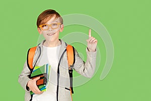 Cute student boy with school bag over green background. Happy kid in glasses pointing up on empty copy space