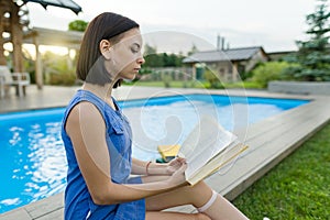 Cute student girl reading book, background swimming pool, lawn near the house. Education, summer, knowledge