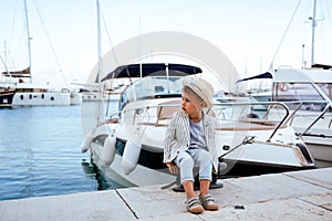 Cute stylish boy sitting on the pier near yacht in marina