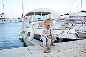 Cute stylish boy sitting on the pier near yacht in marina