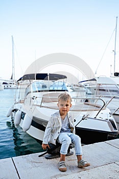 Cute stylish boy sitting on the pier near yacht in marina