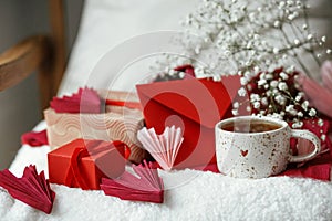 Cute stylish cup of tea with heart on cozy modern armchair with gifts, red envelope and white flowers. Valentine morning surprise