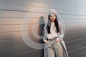 Cute stylish young woman in fashionable knitted hat in long trendy coat in fashionable glasses is standing in the street near gray