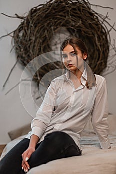 Cute stylish young woman in a fashionable white shirt in vintage black jeans enjoys a rest sitting on the bed in a cozy bedroom.