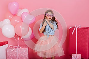 Cute surprised little girl with long brunette hair holding mask on face, looking to camera  on pink background
