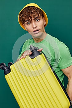 a cute, surprised man stands on a green background, dressed in a yellow panama hat, holding a bright travel suitcase in