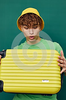 a cute, surprised man stands on a green background, dressed in a yellow panama hat, holding a bright travel suitcase in