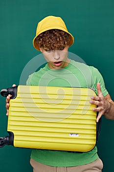 a cute, surprised man stands on a green background, dressed in a yellow panama hat, holding a bright travel suitcase in