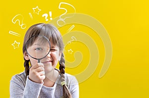 Cute suspicious little girl looking through a magnifying glass on yellow background with copy space