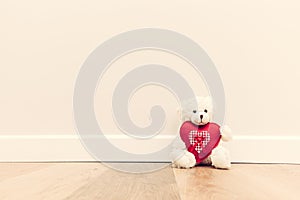 Cute teddy bear with big red plush heart. Sitting on wooden floor against white wall.