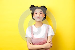 Cute teen asian girl pucker lips for kiss, cross arms on chest and look romantic at camera, yellow background