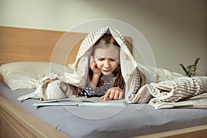 Cute teen child girl lying and reading book on bed under the blanket