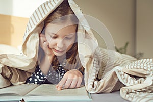 Cute teen child girl lying and reading book on bed under the blanket