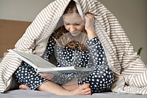 Cute teen child girl sitting and reading book on bed under the blanket