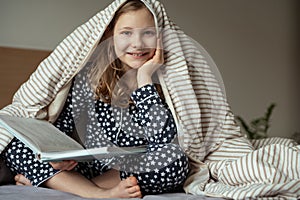 Cute teen child girl sitting and reading book on bed under the blanket