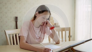 Cute teen girl girl looks at a book and writes in a notebook then looks at her watch. homeschooling concept. Living room in backgr